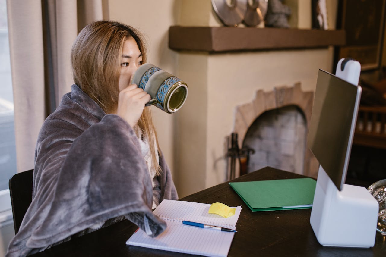 Adult woman sipping coffee while working remotely at home, with a cozy setup.